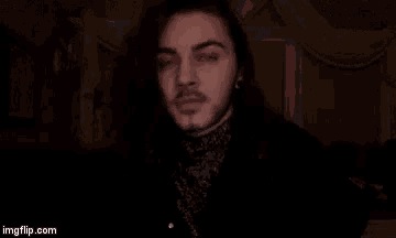 a man with long hair and a beard is standing in a dark room looking at the camera .