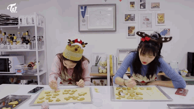 two girls wearing santa hats and reindeer antlers are preparing cookies for christmas