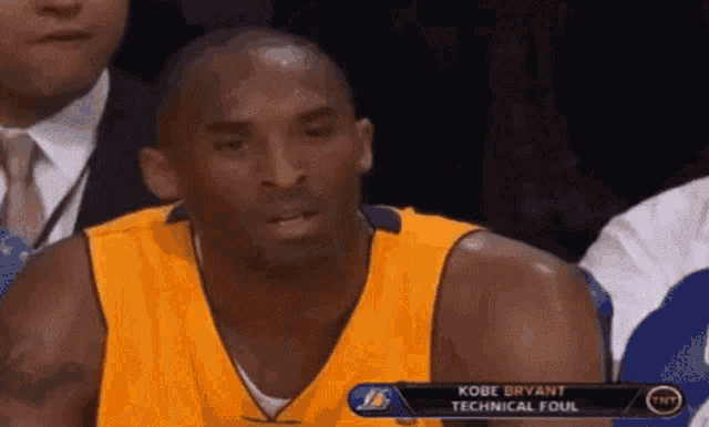 kobe bryant is sitting in the stands during a basketball game with a technical foul on the screen behind him .