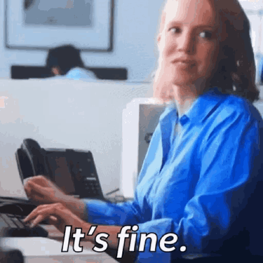 a woman in a blue shirt is sitting at a desk typing on a keyboard and says it 's fine .