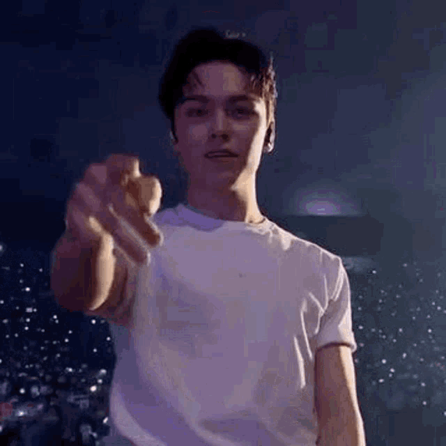 a young man in a white t-shirt is pointing his finger at the camera while standing on a stage .