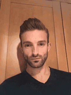 a man with a beard is standing in front of a wooden wall and looking at the camera .