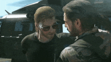 a man wearing sunglasses talks to another man in front of a helicopter