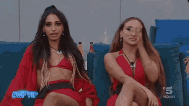 two women are sitting next to each other on a couch and one of them is wearing a red bikini top .
