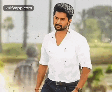 a man with a beard is wearing a white shirt and jeans and standing in a park .