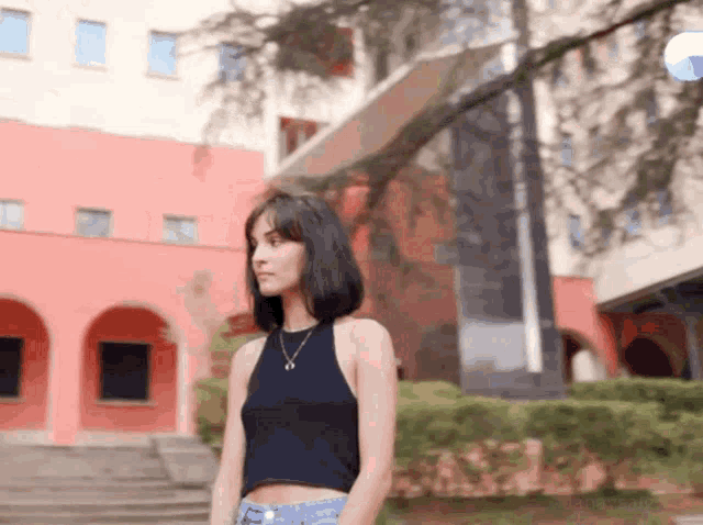 a woman in a black tank top and blue jeans stands in front of a building