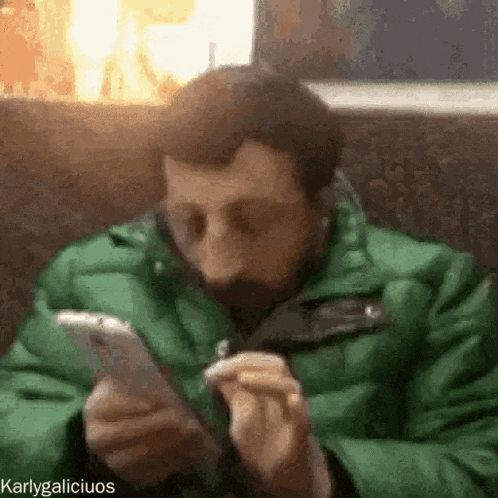 a man in a green jacket is smoking a cigarette and looking at his phone .