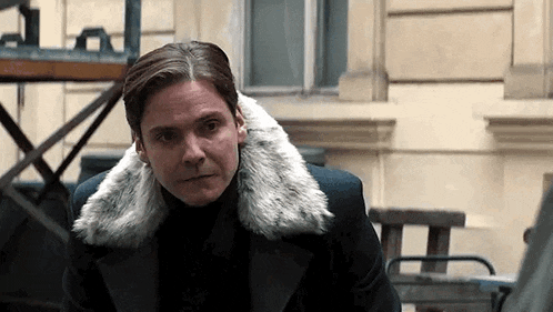 a man in a fur coat is standing in front of a building and looking at the camera .