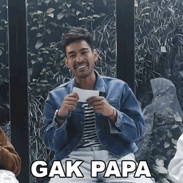 a man in a denim jacket is smiling while holding a piece of paper with the words " gak papa " on it .