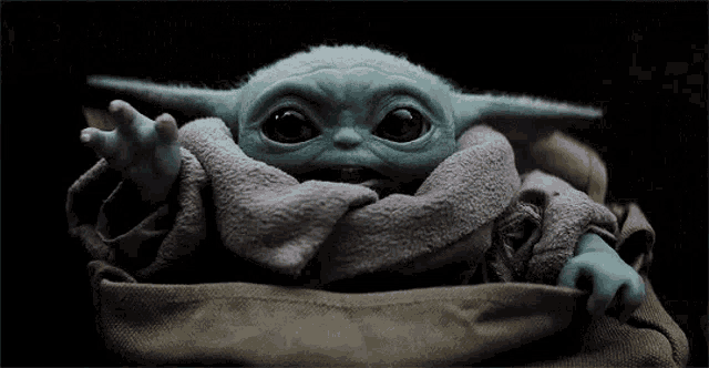 a baby yoda toy is sitting in a blanket and looking at the camera .