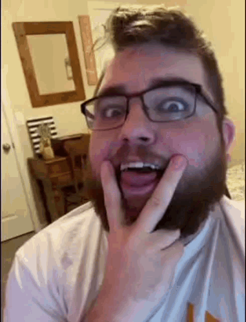 a man with a beard and glasses is making a funny face with his hands on his face .