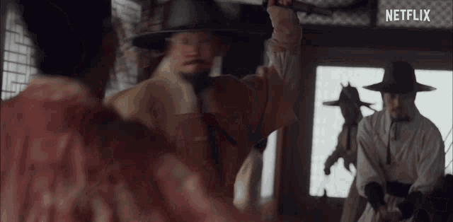 a man in a hat is holding a sword in his hand in a room with other men .