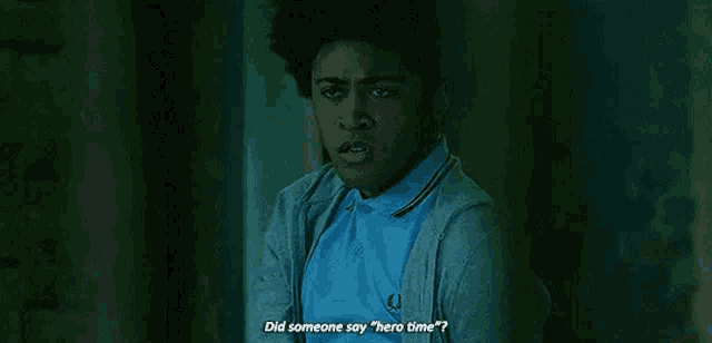 a young man in a blue shirt and cardigan is standing in a dark room and talking to someone .