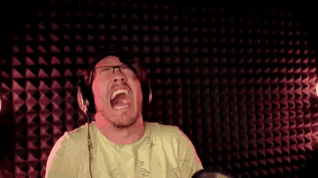 a man wearing headphones and glasses is laughing in front of a microphone in a recording studio .