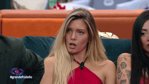 a woman in a red dress sits on a couch in front of a sign that says #grandefratello
