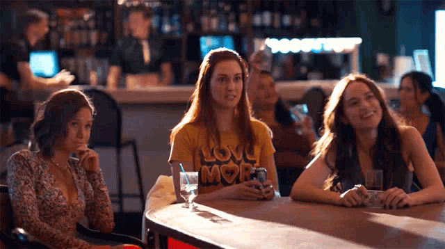 three women are sitting at a table with one wearing a yellow shirt that says love more