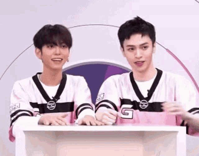 two young men are standing next to each other at a table wearing pink jerseys .