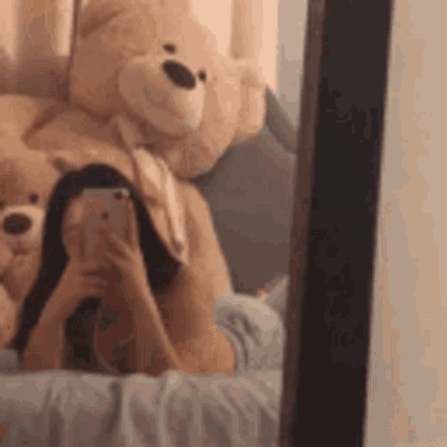 a woman is taking a selfie in front of a mirror with a teddy bear in the background .