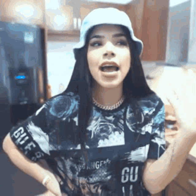 a girl wearing a hat and a t-shirt is standing in a kitchen .