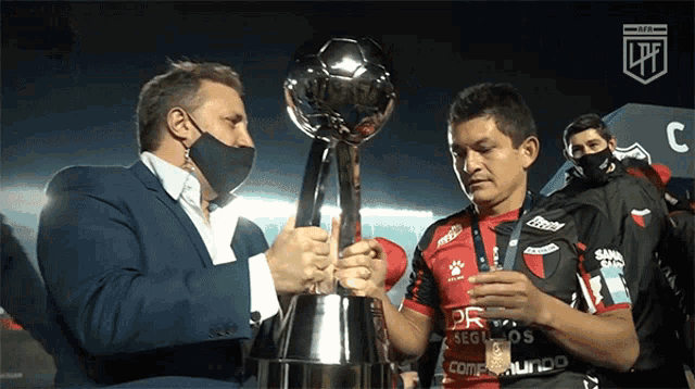 a man holding a trophy next to another man wearing a jersey that says seguros