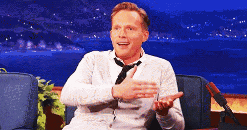 a man in a white shirt and tie is sitting in front of a microphone and making a funny face .
