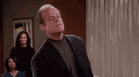 a man in a suit and turtleneck is dancing in a room with a woman in the background .