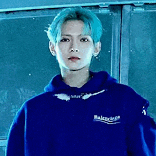 a young man with blue hair is wearing a blue hoodie and standing in front of a window .