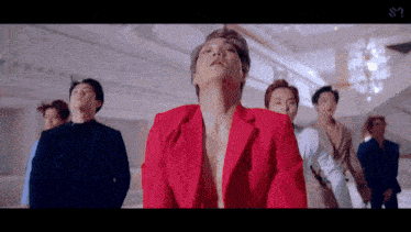 a group of men are standing in a room and one is wearing a red jacket