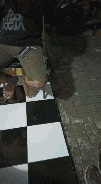 a man wearing a black shirt with the word iggm on it sits on a checkered tile floor
