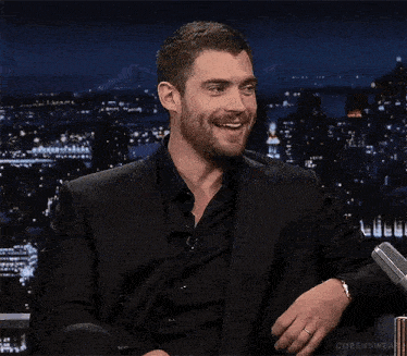 a man in a suit and black shirt is laughing while sitting in front of a city skyline .