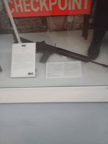 a gun in a glass case with a checkpoint sign behind it