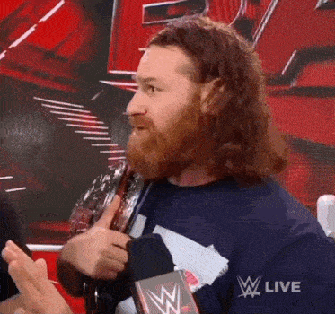 a man with a beard is talking into a microphone while holding a wrestling championship belt .
