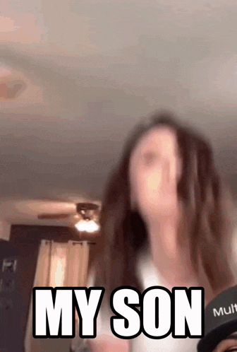 a woman is standing in a living room with the words `` my son '' written on the screen behind her .