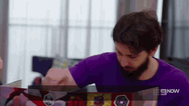 a man in a purple shirt sits at a table with a box in front of him that says 9 now on it