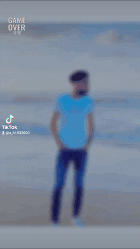 a man in a blue shirt stands on a beach with the words game over behind him