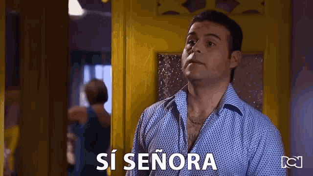 a man in a blue shirt is standing in front of a yellow door and says si señora