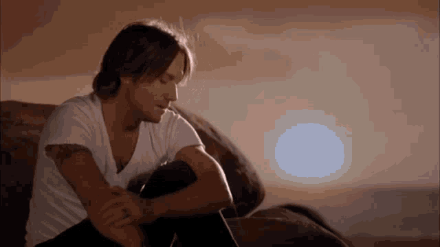 a man in a white shirt is sitting on a rock looking at the sun .