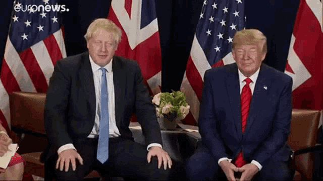 two men in suits and ties are sitting next to each other in front of american and british flags .