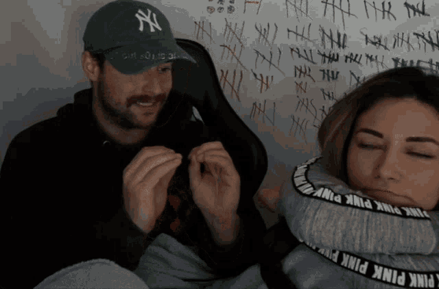 a man wearing a ny hat sits next to a woman wearing pink pink