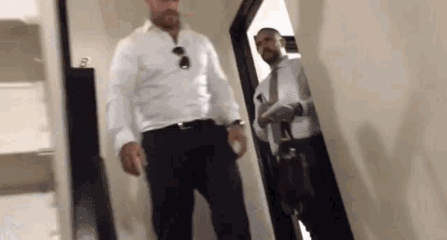a man in a white shirt and black pants is standing in a hallway next to another man in a suit and tie .