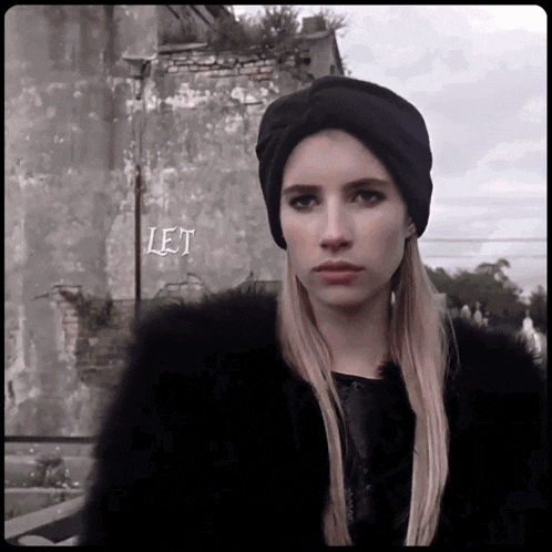 a woman wearing a black hat and a fur coat stands in front of a wall that says let