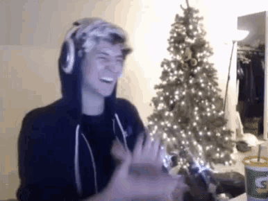 a man wearing headphones and a hoodie is laughing in front of a christmas tree .