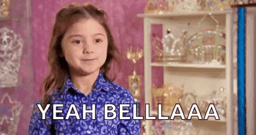 a little girl in a blue shirt is standing in front of a shelf and says `` yeah bellaa '' .