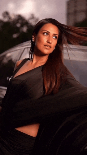 a woman in a black dress is standing in front of a car with her hair blowing in the wind