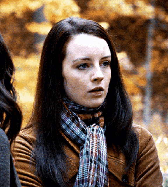 a woman wearing a plaid scarf and a brown jacket has a serious look on her face