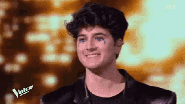 a young man with blue tears on his face is smiling and wearing a black jacket with the word voice on the bottom