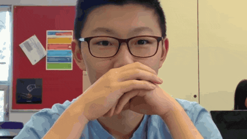 a boy with glasses covering his mouth with his hands in front of a red board with sticky notes on it