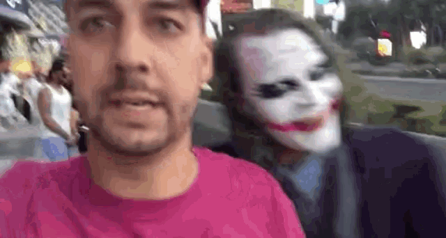 a man in a pink shirt is taking a selfie with a joker mask behind him .