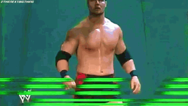 a man without a shirt is standing in front of a green background with a wrestling match in progress .