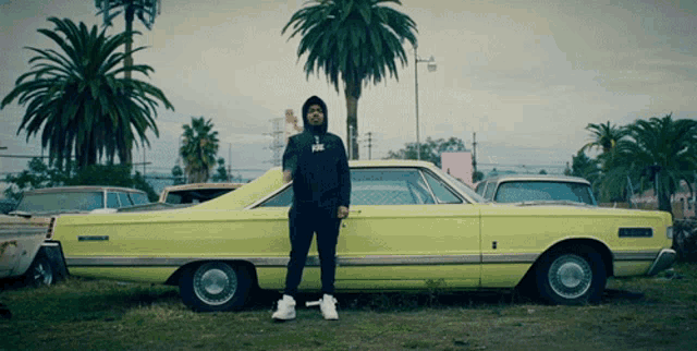 a man in a black hoodie stands in front of a yellow car